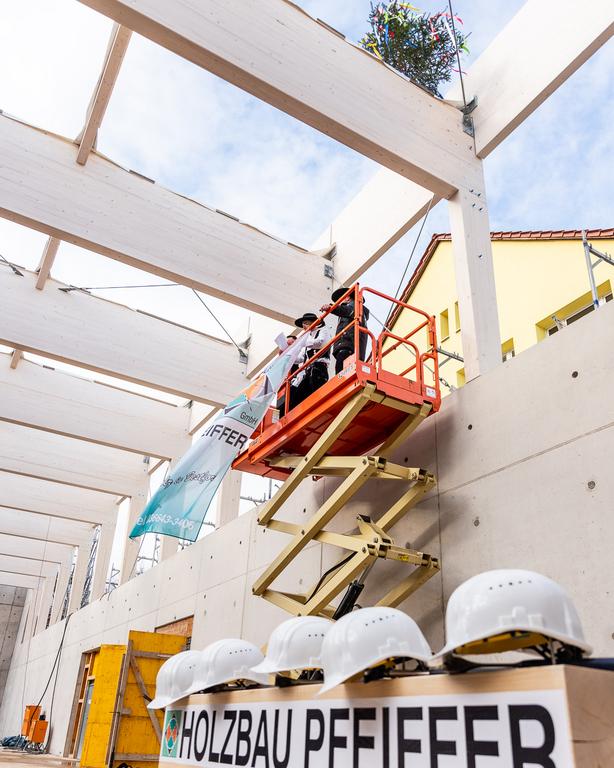 Zwei Personen in Anzug und Hut stehen auf einer Hebebühne in einer Rohbauhalle mit sichtbaren Holzbalken und Richtkranz. Im Vordergrund mehrere weiße Bauhelme auf einem Schild mit der Aufschrift 'HOLZBAU PFEIFFER'.