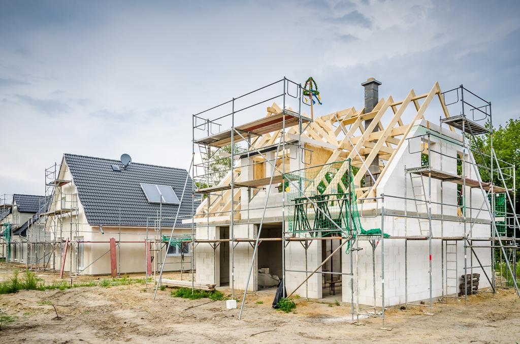 Rohbau eines Hauses mit sichtbarem Dachstuhl und Gerüst, daneben ein fertiges Haus mit Solarpanelen auf dem Dach.