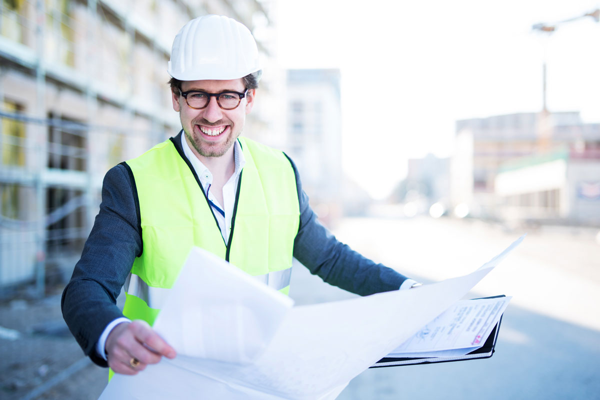 Bauleiter steht mit Helm und Warmweste auf Baustelle und ließt Baupläne