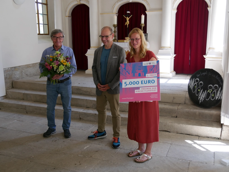 Staatssekrektär Torsten Weil überreicht einen symolischen Gewinnerscheck und Blumen an die Gewinner in der Kirche