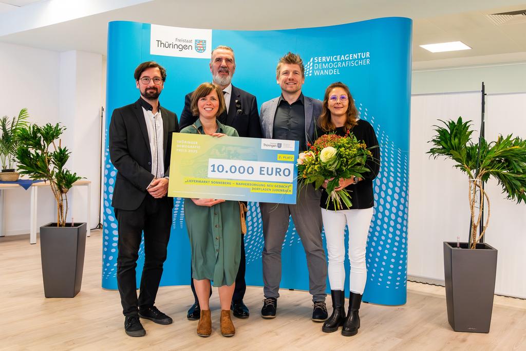 Fünf Personen stehen vor einem blauen Hintergrund mit Logos, eine hält einen großen Scheck über 10.000 Euro, eine weitere einen Blumenstrauß.
