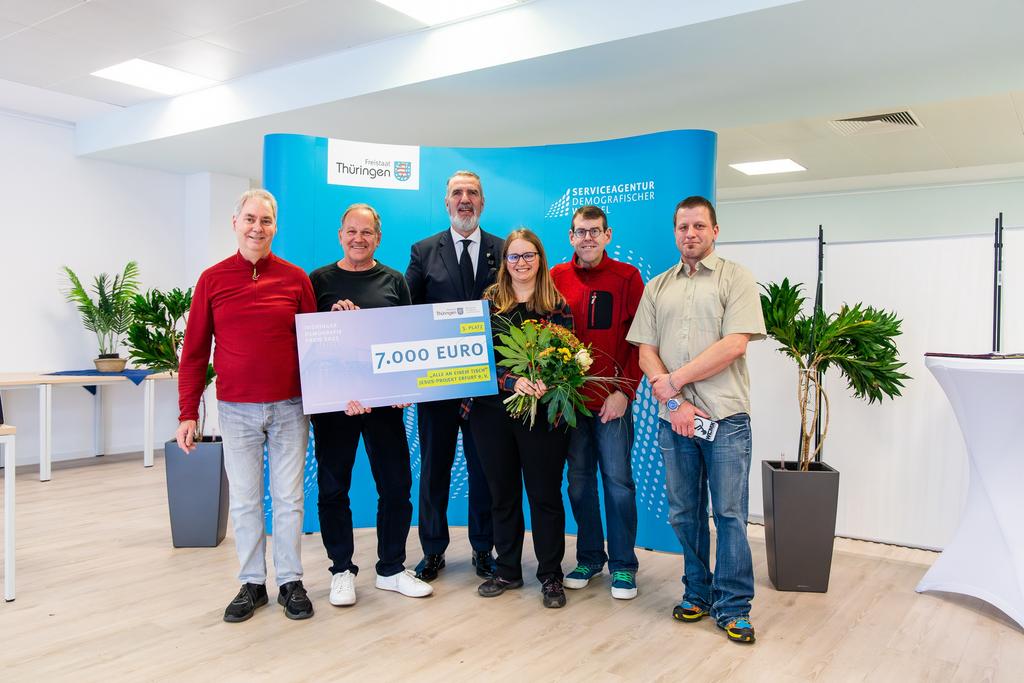 Sechs Personen stehen vor einem blauen Hintergrund mit Text, zwei halten einen großen Scheck, zwei halten Blumensträuße, Raum mit Pflanzen und Stehtischen.