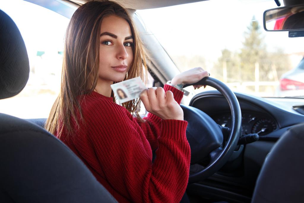 Ein junges Mädchen sitzt am Fahrzeugsteuer und hält einen Führerschein in der Hand.