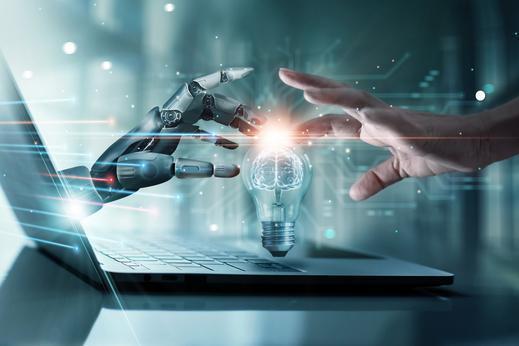 Roboterhand und menschliche Hand berühren sich über einer Laptop-Tastatur, zwischen ihnen schwebt eine leuchtende Glühbirne.