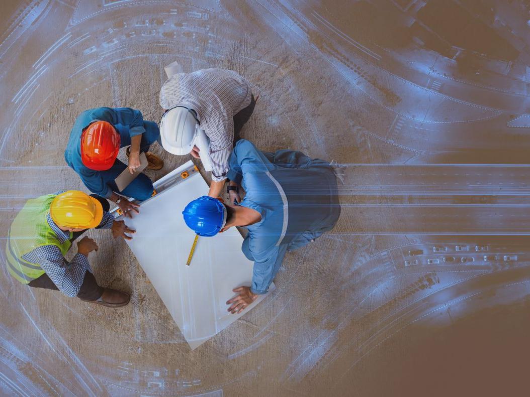 Symbolisches Bild: Vier Ingenieure mit Bauarbeiterhelmen neigen sich über Plan zu Straßenbauprojekt