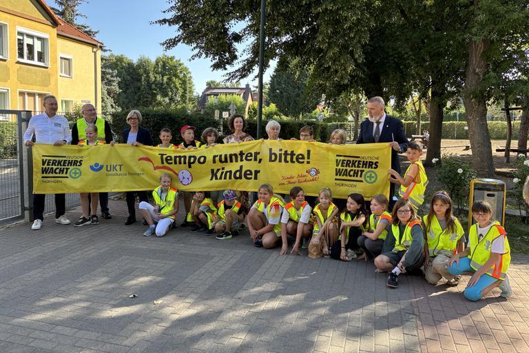 Kinder in gelben Warnwesten sitzen und stehen vor einem Baum, halten ein gelbes Banner mit der Aufschrift 'Tempo runter, bitte! Kinder' und Logos von Verkehrswacht und UKT.