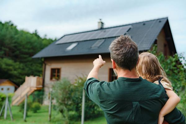 Mann mit kleinem Kind auf dem Arm steht im Garten und zeigt auf die Photovoltaikanlage seines Hauses