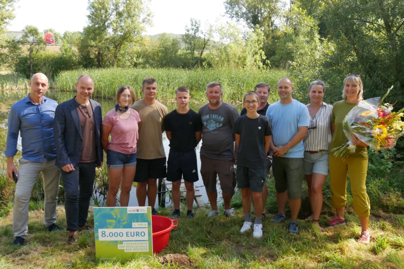 Ein Gruppe Menschen steht vor dem Teich. Staatssekretär Torsten Weil überreicht den Gewinnern einen symbolischen Scheck