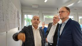 Drei Männer mit weißen Hemden und dunklen Anzügen stehen in einem Raum. An der Wand hängen Baupläne. Diese werden von dem Projektleiter den anderen zwei Herren erläutert.