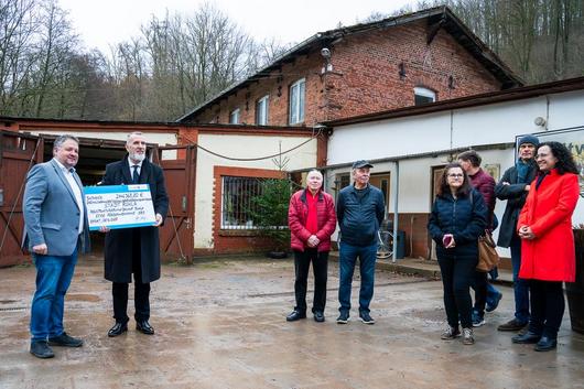Minister Steffen Schütz steht vor einem alten Gebäude am Bauhof Ruhla und überreicht einen symbolischen Fördermittelbescheid an den Bürgermeister. Einige Bürger stehen dabei und freuen sich