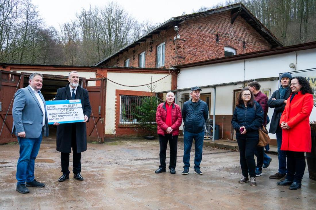 Minister Steffen Schütz steht vor einem alten Gebäude am Bauhof Ruhla und überreicht einen symbolischen Fördermittelbescheid an den Bürgermeister. Einige Bürger stehen dabei und freuen sich