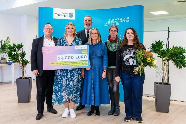 Sechs Personen stehen vor einem blauen Hintergrund mit Text, eine Frau hält einen großen Scheck, eine hält einen Blumensträuße, Raum mit Pflanzen und Stehtischen.