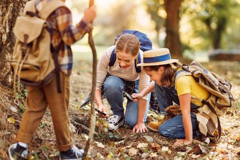 Zwei Kinder, Junge und Mädchen mit Rucksäcken, untersuchen die Umgebung durch die Lupe, während sie an einem sonnigen Tag während des Ökologie-Schulunterrichts im Freien die Natur des Waldes erkunden