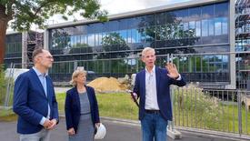 Eine Frau und zwei Männer stehen vor einem Bauzaum. Hinter dem Bauzaum ist eine Wiese mit einem Sandhaufen. Dahinter ist ein großes langezogenes zweietagiges moderen Gebäude mit Glasfronten. Die Frau hält einen weißen Bauhelm in der Hand. Der Mann links neben ihr erklärt mit zeigender Hand etwas zum Baugeschehen.
