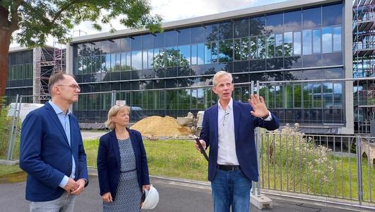 Eine Frau und zwei Männer stehen vor einem Bauzaum. Hinter dem Bauzaum ist eine Wiese mit einem Sandhaufen. Dahinter ist ein großes langezogenes zweietagiges moderen Gebäude mit Glasfronten. Die Frau hält einen weißen Bauhelm in der Hand. Der Mann links neben ihr erklärt mit zeigender Hand etwas zum Baugeschehen.