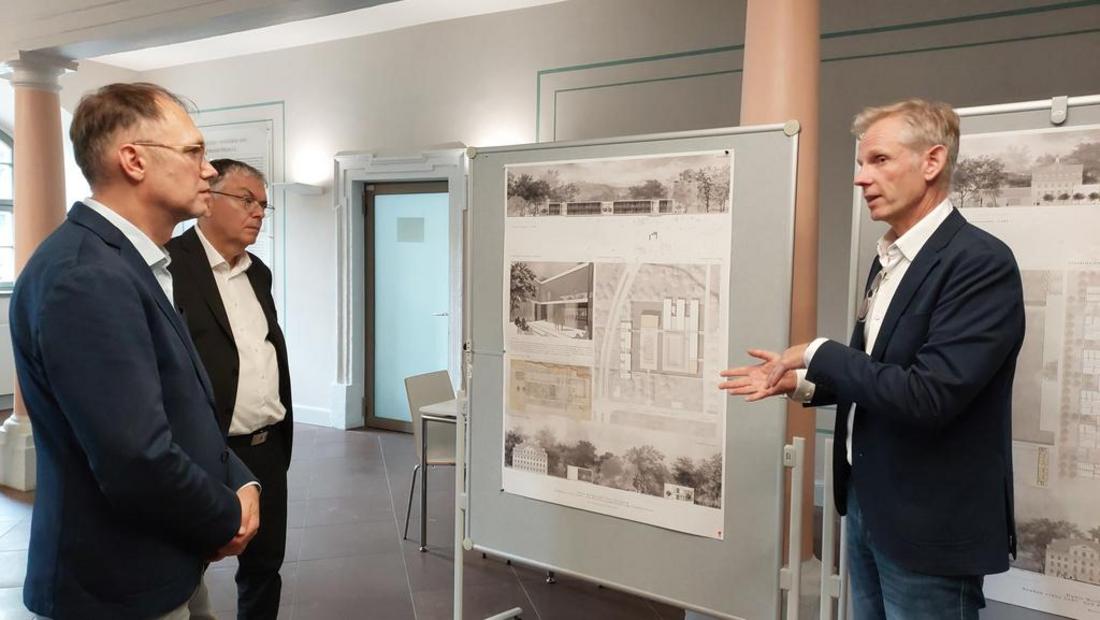 Drei Männer mit weißen Hemden und dunklen Anzügen stehen in einem Raum vor einer grauen Pinnwand. Auf der Pinnwand ist ein Architekturenplan vom Erweiterungsbau der Dualen Hochschule Gera-Eisenach angebracht.