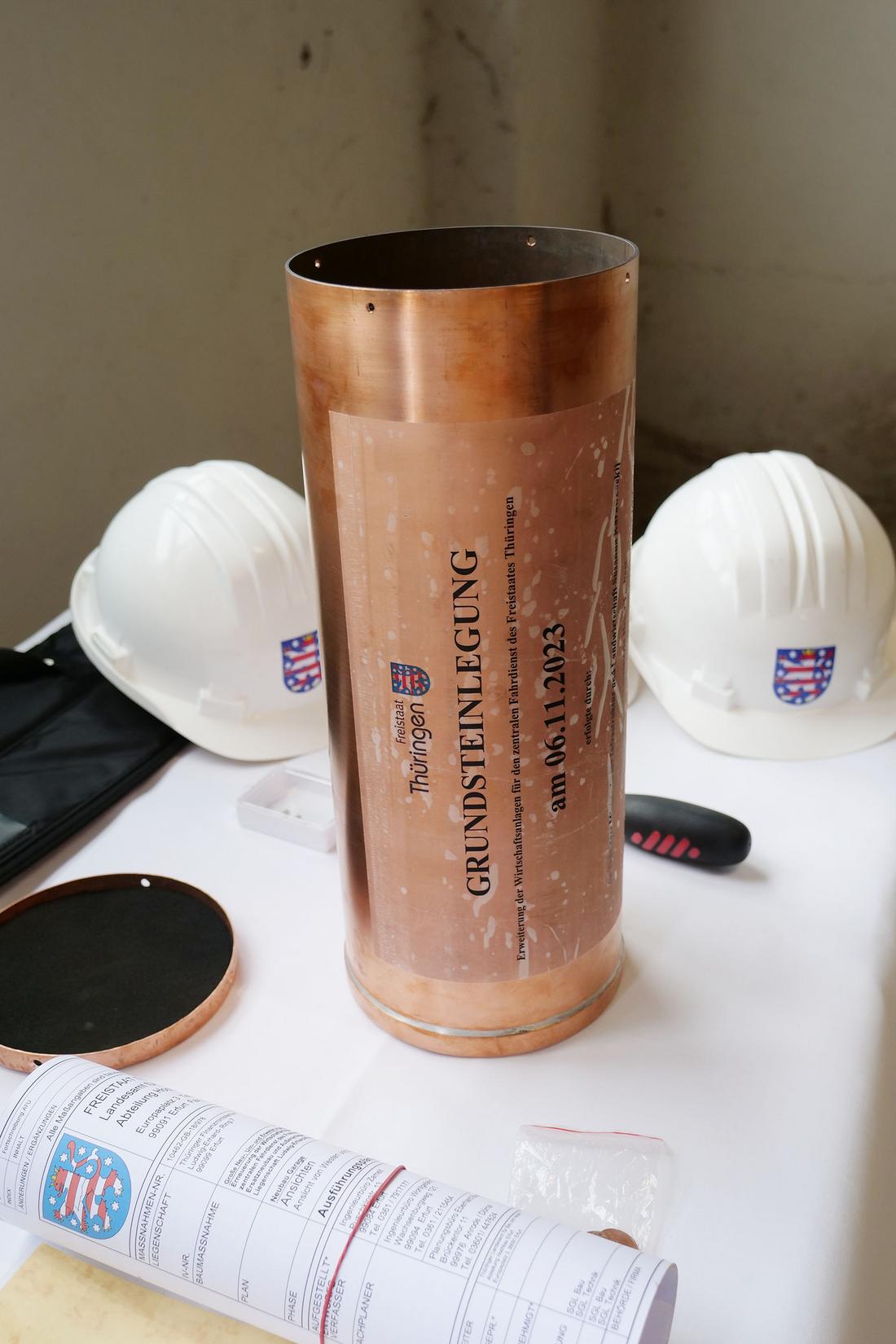 Tisch mit Bauunterlagen, zwei Bauhelme im Hintergrund und in der Mitte eine Zeitkapsel für die Grundsteinlegung