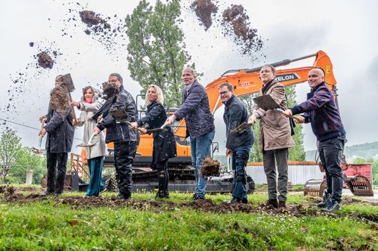 Ministerin Karawanskij und weitere Personen werfen mit dem Spaten Erde in die Höhe, im Hintergrund steht ein oranger Bagger