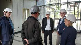 Eine Frau und vier Männer stehen in einem im Rohbau befindlichen Gebäude im Kreis und unterhalten sich. Alles haben weiße Bauhelme auf.