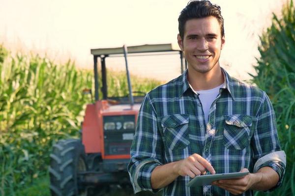 Portrait eines jungen Bauern und Traktor auf dem Maisfeld mit Laptop in der Hand