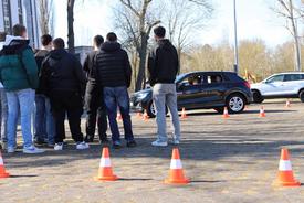 Mehrere Jugendliche stehen aus dem Schulhof und verfolgen das Einparken eines PKW auf einem markierten Parkplatz durch einen Mitschüler.