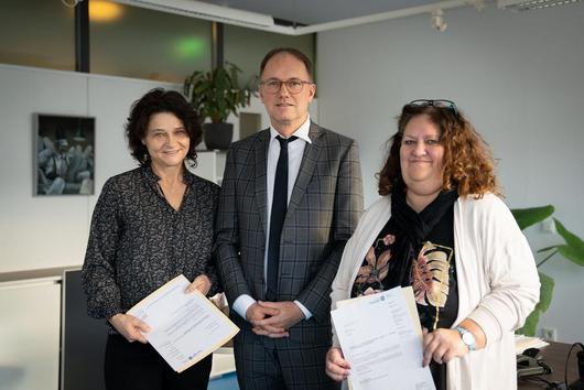 drei Personen, in der Mitte Staatssekretär Dr. Knoblich, stehen nebeneinander. Die beiden Frauen halten Ihre Mitteilungen über ihre Wahl zur Gleichstellungsbeauftragten und stellvertretenden Gleichstellungsbeauftragten in den Händen