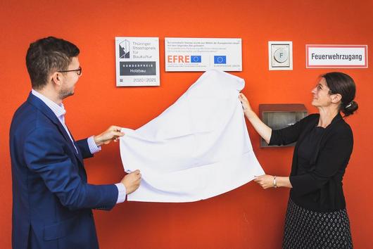 Staatsekretärin Schönig und der Ilmenauer Oberbürgermeister enthüllen die an der Wand angebrachte Plakette des Thüringer Staatspreises