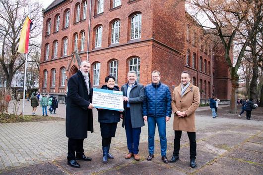 Minister Steffen Schütz steht gemeinsam mit drei weiteren Herren und der Schulleiterin der Martinischule vor dem alten Klinker Schulgebäude und überreicht einen Fördermittelscheck über 6 Millionen Euro