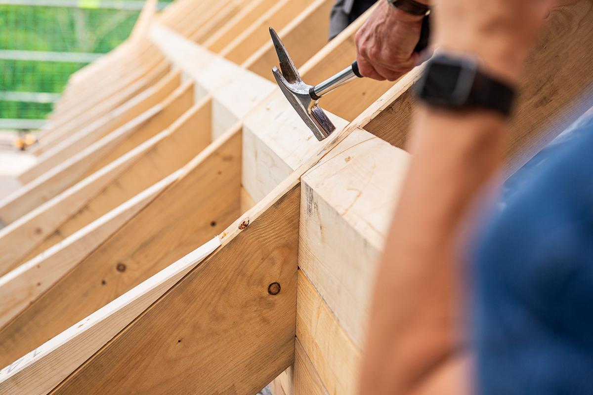 Hand hält Hammer und schlägt Nagel in Holzbalken des Dachstuhls bei Richtfest.