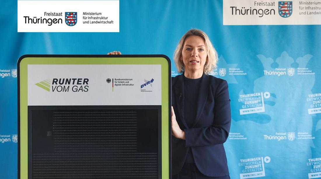 Ministerin Karawanskij steht mit Dialogdisplay vor blauem Banner des TMIL