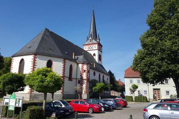 Große Kirche von Herbsleben, im Vordergrund ein Parkplatz mit Fahrzeugen, rechts ein Laubbaum, blauer Himmel