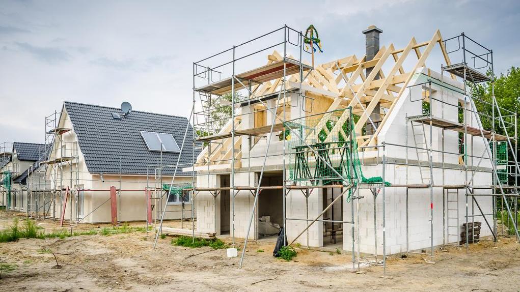 Rohbau eines Hauses mit sichtbarem Dachstuhl und Gerüst, daneben ein fertiges Haus mit Solarpanelen auf dem Dach.