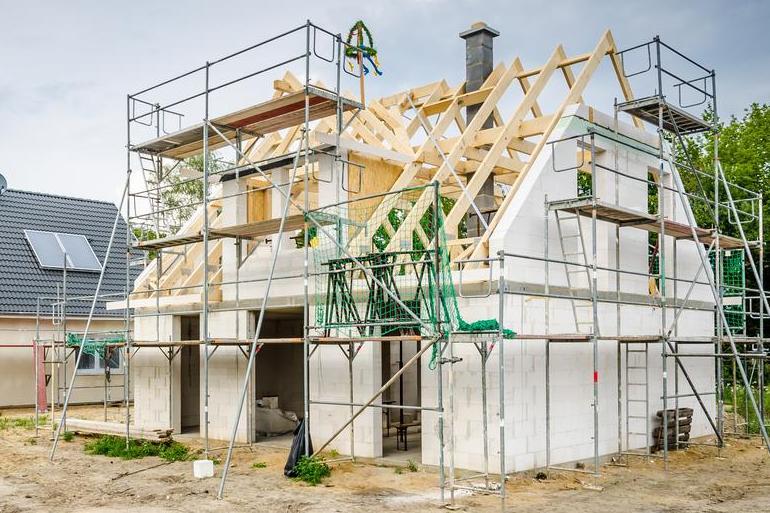 Rohbau eines Hauses mit sichtbarem Dachstuhl und Gerüst, daneben ein fertiges Haus mit Solarpanelen auf dem Dach.